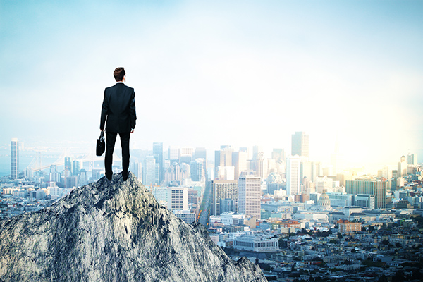 Leadership concept Back view of young man in suit and with briefcase in hand standing on mountain top. City background with copy space. Leadership concept